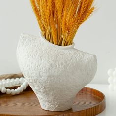 a white vase with some yellow grass in it on a wooden tray next to beads