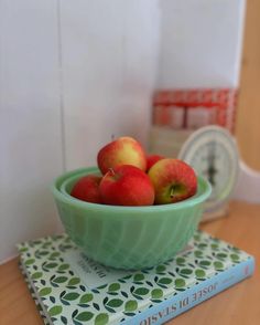 a green bowl filled with apples sitting on top of a book next to a clock