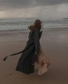 a woman is walking on the beach with her dress blowing in the wind and dark clouds