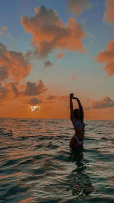 a woman standing in the ocean at sunset raising her arms up to the sun above water