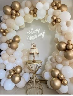 a white and gold birthday cake surrounded by balloons in the shape of a circle on a table