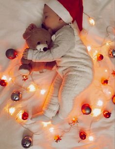 a baby in a santa hat sleeping on a blanket with christmas lights around it and a teddy bear