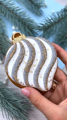 a hand holding a decorated christmas ornament in front of a pine tree with gold and white stripes