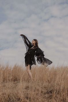 a woman standing in a field with her arms spread out to the side, wearing a black dress