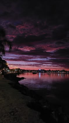 the sky is purple and pink as people walk on the shore at night near water