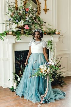 a woman standing in front of a fire place holding a bouquet and wearing a blue dress