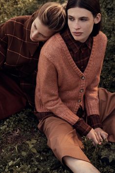 two women are sitting on the ground in front of some bushes and grass, one is wearing a brown cardigan