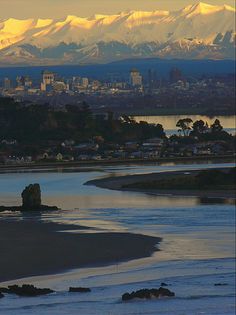 the mountains are covered in snow as seen from across the river, with houses and buildings on either side