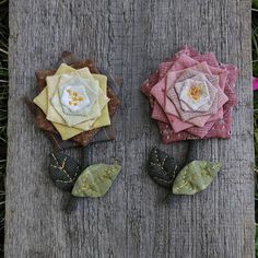 two fabric flowers sitting on top of a piece of wood next to each other with leaves