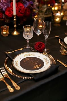 a table set with gold and black place settings, silverware and red rose centerpieces