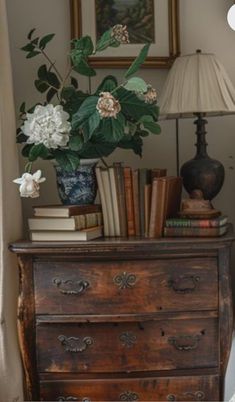 a dresser with books and flowers on it in a room next to a lamp, framed by a window