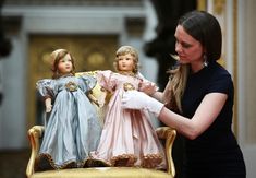 a woman in black shirt and white gloves with two dolls on gold chair next to each other