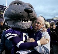 two men hugging each other in front of a mascot at a football game with people watching from the bleachers