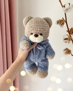 a hand holding a crocheted teddy bear in front of a window with pink curtains