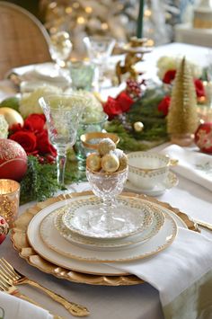 the table is set for christmas dinner with gold and white plates, silverware, and red roses