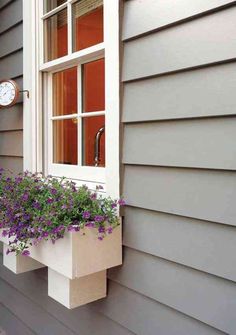 a window box filled with purple flowers next to a gray wall and clock on the side of a house