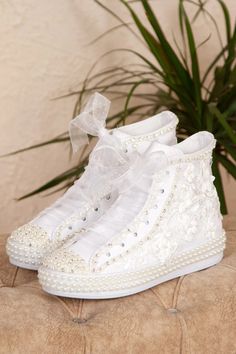 a pair of white high top sneakers with pearls and bows on the side, next to a potted plant