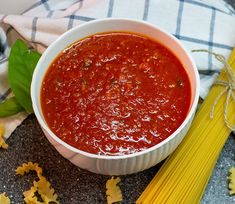 a bowl of tomato sauce next to some pasta and leaves on a tablecloth with a blue checkered towel in the background