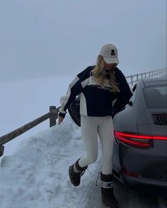 a woman in white pants and black jacket standing next to a car on snow covered ground