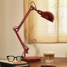 a red desk lamp sitting on top of a wooden table next to a cup of coffee