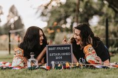 two women sitting on the grass holding bottles and posing for a photo with a sign that says best friends established