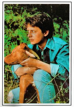 a man holding a brown dog in his lap while sitting on the ground next to tall grass