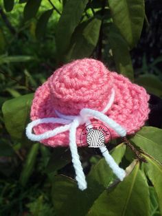 a pink crocheted hat hanging from a tree branch with leaves around it and a white ribbon tied to the brim