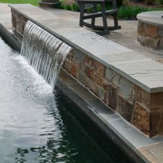 a small waterfall in the middle of a pond with benches around it and an empty park bench on the other side