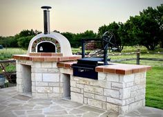 an outdoor pizza oven built into the side of a stone patio with grass and trees in the background