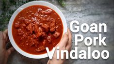 two hands holding a white bowl filled with meat and tomato sauce that says goan pork vindalloo