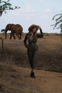 a woman in a suit and hat walking down a dirt road next to an elephant