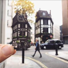 a person holding up a cut out of a building on the street with cars passing by