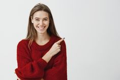 a smiling woman in a red sweater points to the side with her index finger and smiles at the camera