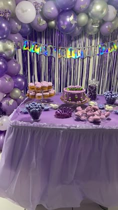 a table topped with purple and white desserts next to lots of balloon garlands