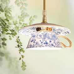 a blue and white flowered lamp hanging from a gold colored light fixture with green leaves in the background