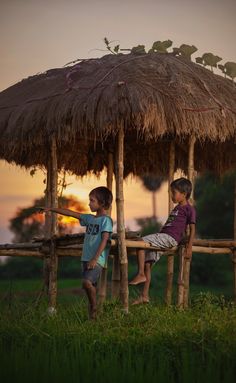 two young boys sitting on a wooden bench under a thatch roof at sunset with the sun setting behind them