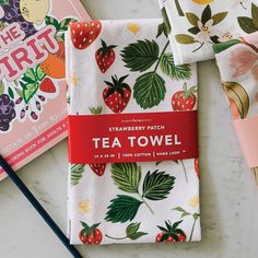 two tea towels with strawberries on them next to a notebook and pencil, which are also in front of the strawberry match book