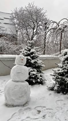 a snowman sitting next to two small trees covered in snow on a snowy day