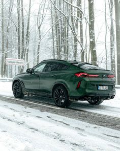 a green car driving down a snow covered road next to trees in the wintertime