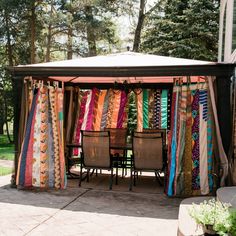 a group of chairs sitting under a tent covered in colorful blankets and scarves with trees in the background