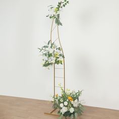 a plant stand with flowers and greenery in it on a wooden floor next to a white wall