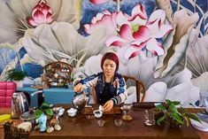 a woman sitting at a table with tea cups in front of her and flowers on the wall behind her