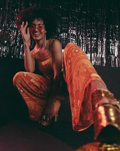 a woman sitting on the ground with her hand up to her face and wearing an orange dress