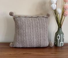 a knitted pillow with pom poms sits next to a vase filled with flowers