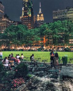 people are sitting at tables in the middle of a park with tall buildings behind them