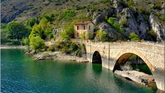 an old stone bridge over a body of water with a house on the cliff in the background