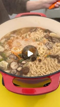 a red pot filled with noodles and mushrooms on top of a yellow countertop next to a person holding a carrot