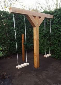 a wooden swing set with two swings attached to it and some bushes in the background