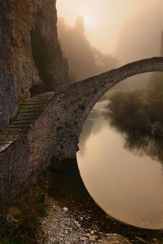 an old stone bridge over a river in the middle of mountains and foggy sky