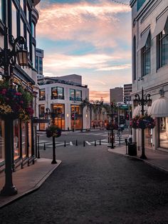 an empty city street at dusk with the sun going down in the distance and flowers hanging on the lampposts